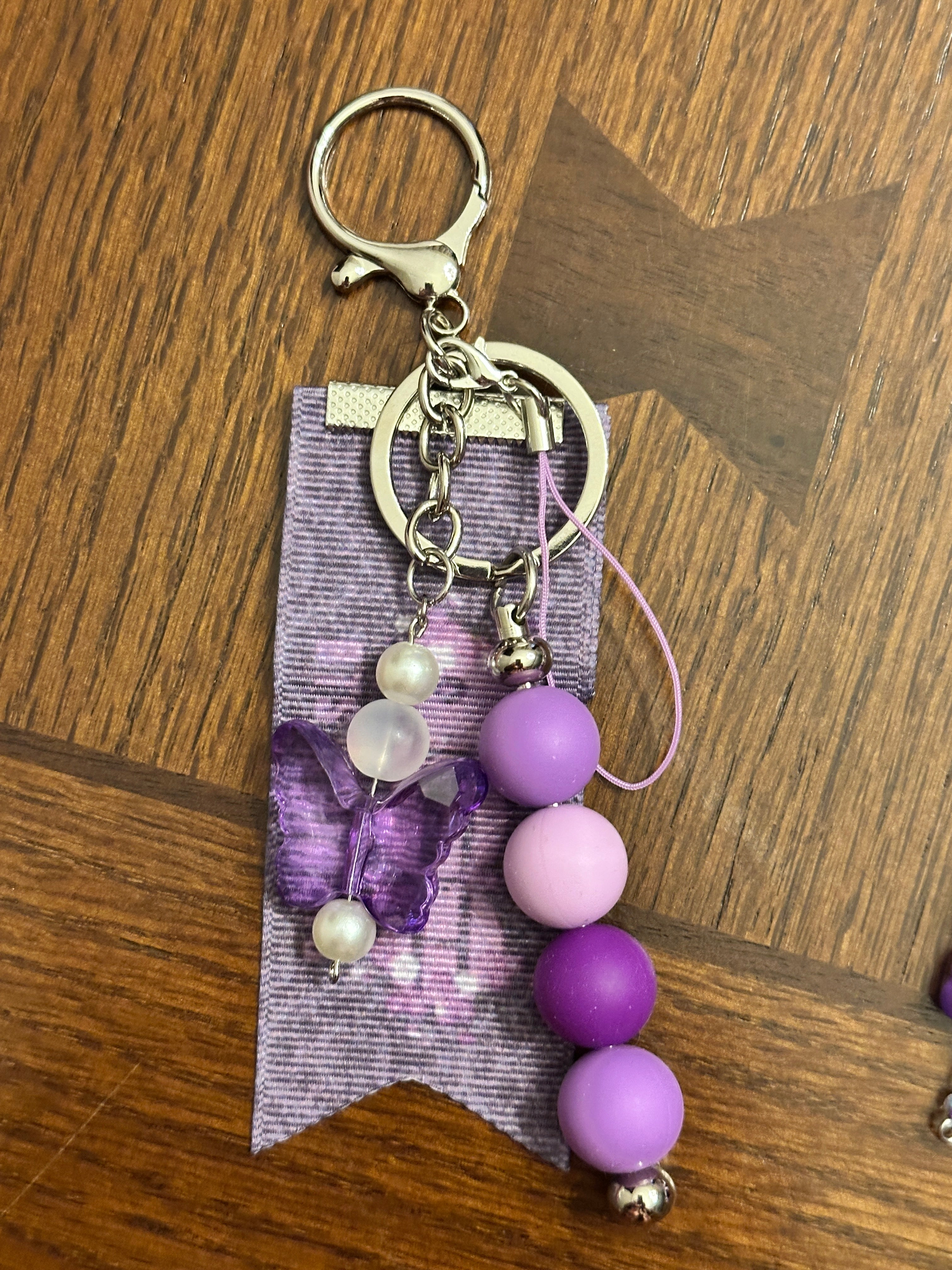 Handcrafted BTS-inspired purple keychain with layered purple beads, butterfly charm, pearl accents, and custom-designed purple ribbon tag by FyaStudios on silver lobster clasp hardware.